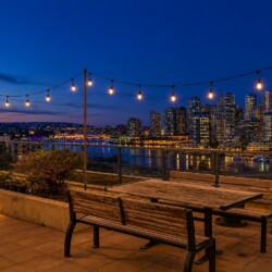 Rooftop patio at dusk with string lights and wooden table and bench, overlooking a city skyline across the water.