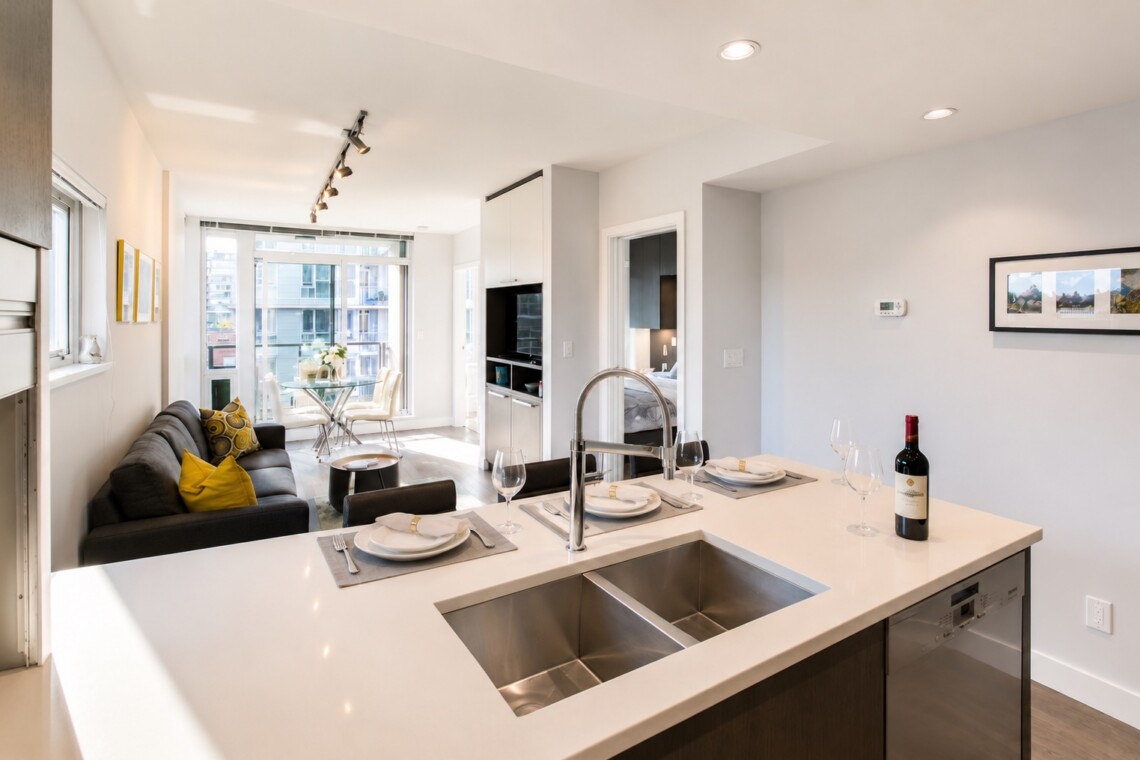 Open-concept kitchen and living area with a white island, sink, wine bottle, and a table-setting by large windows overlooking the city.
