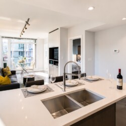 Open-concept kitchen and living area with a white island, sink, wine bottle, and a table-setting by large windows overlooking the city.