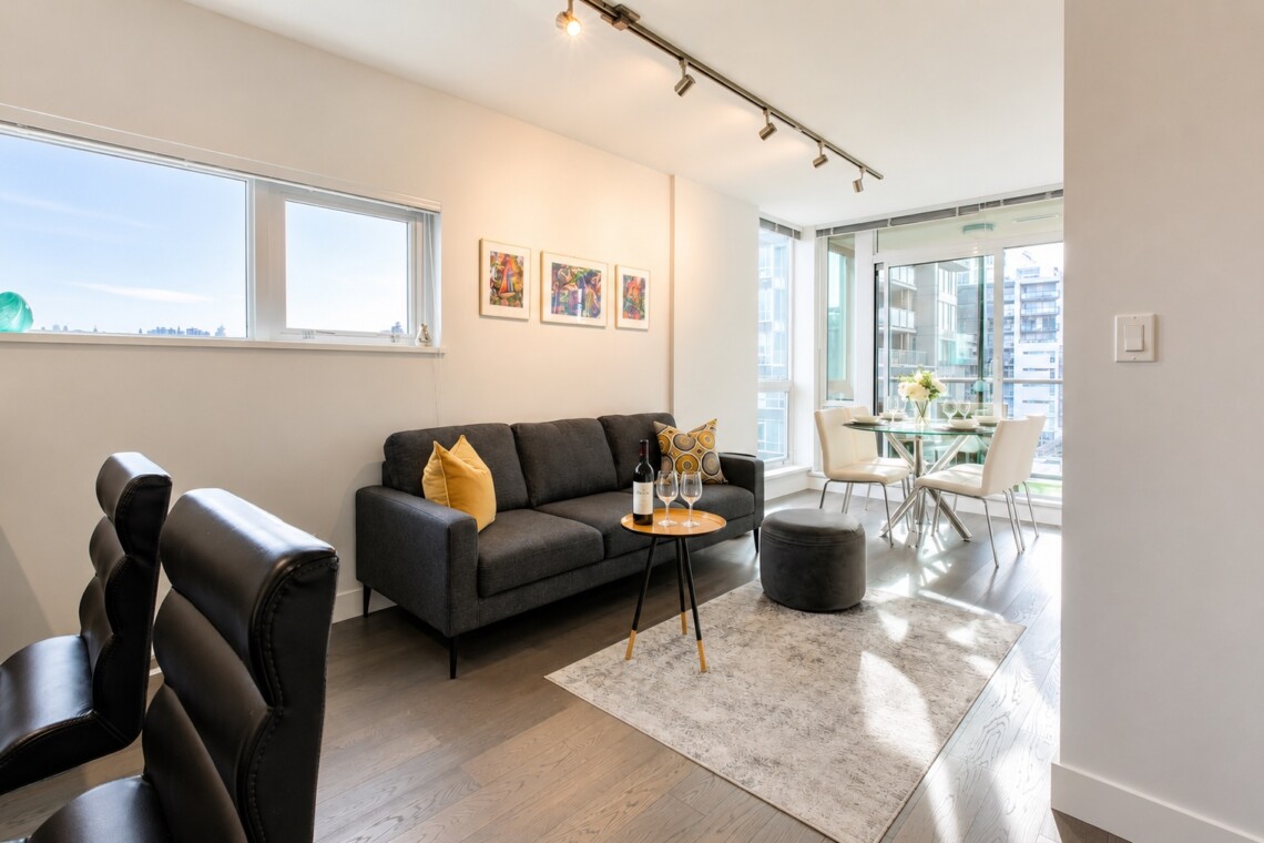 Open living area with a dark gray sofa, yellow cushions, and a round coffee table holding a wine bottle and glasses near a dining nook by large windows.