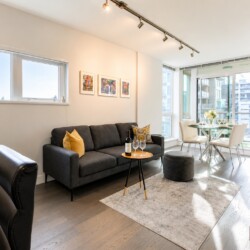 Open living area with a dark gray sofa, yellow cushions, and a round coffee table holding a wine bottle and glasses near a dining nook by large windows.