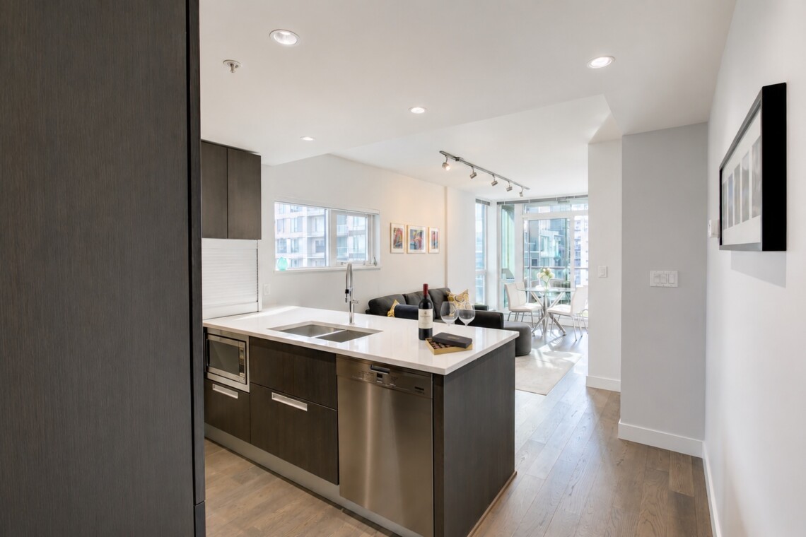 Open-concept kitchen with dark wood cabinets, island sink, stainless appliances, and adjacent living area with a sofa and large windows overlooking the city.