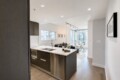 Open-concept kitchen with dark wood cabinets, island sink, stainless appliances, and adjacent living area with a sofa and large windows overlooking the city.