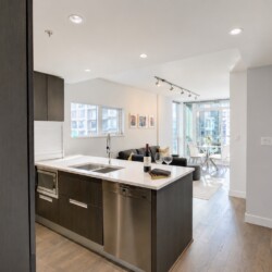 Open-concept kitchen with dark wood cabinets, island sink, stainless appliances, and adjacent living area with a sofa and large windows overlooking the city.