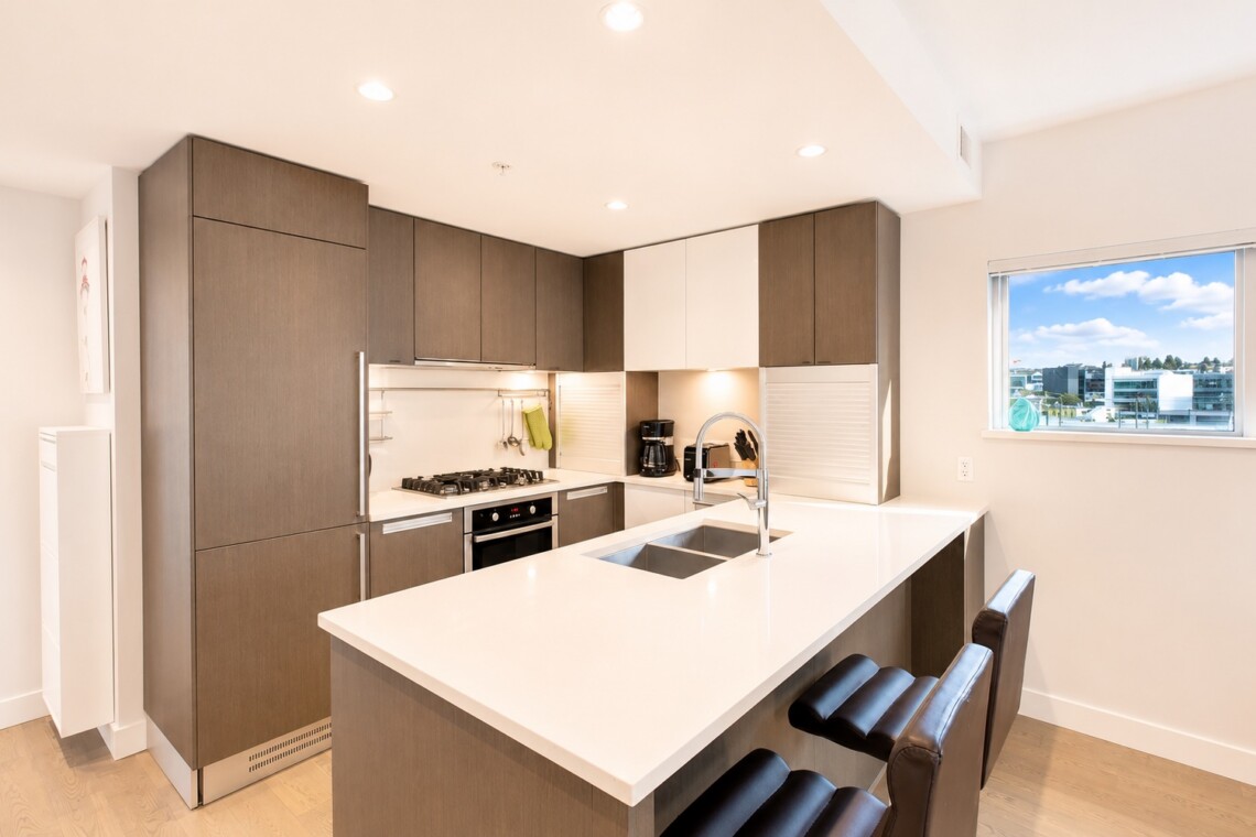 Modern kitchen with white island, double sink, and gray cabinets; large window shows a cityscape outside.