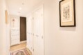 Bright, narrow hallway with white closet doors along the right wall, a white chest of drawers at the far end, and framed art on the walls; a patterned rug on a light wood floor.