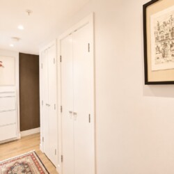 Bright, narrow hallway with white closet doors along the right wall, a white chest of drawers at the far end, and framed art on the walls; a patterned rug on a light wood floor.