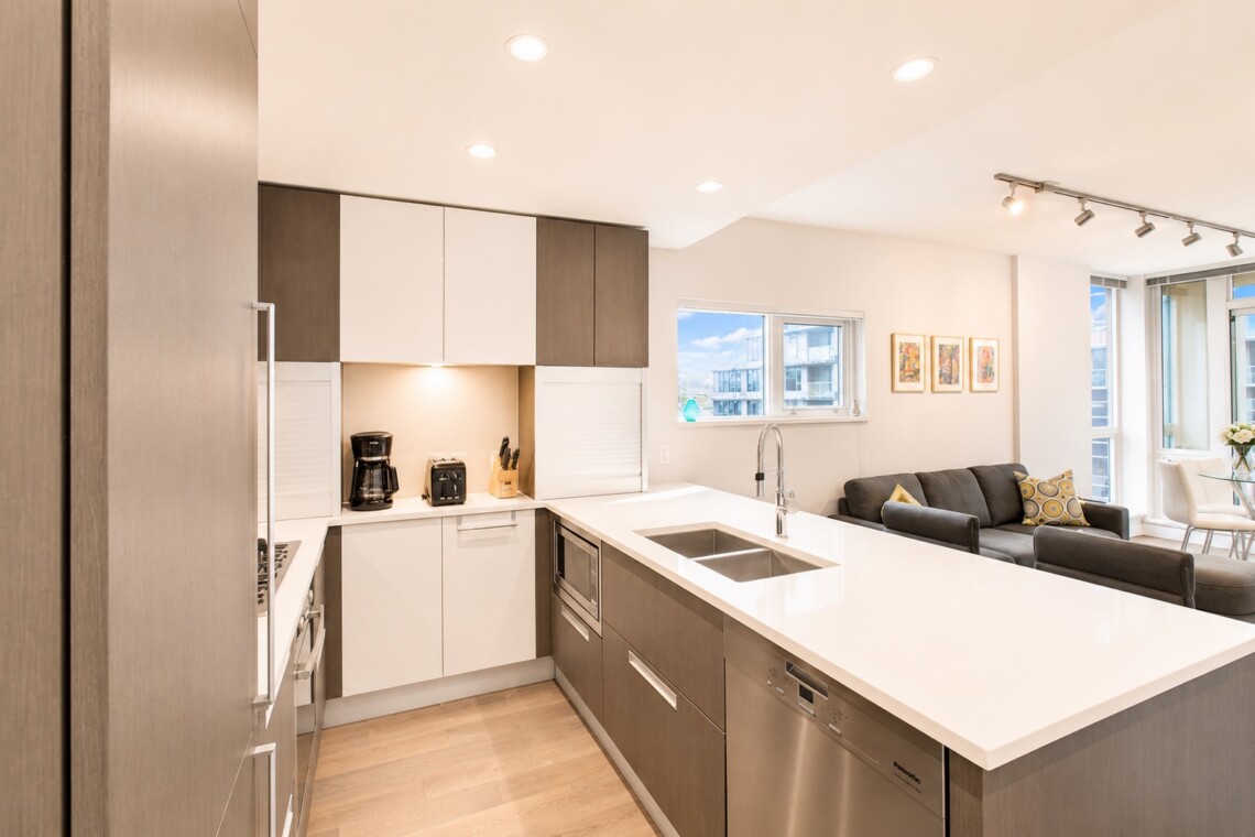 Open-concept kitchen with white and gray cabinets, an island sink, and stainless appliances adjoining a living room with a gray sofa and framed art.