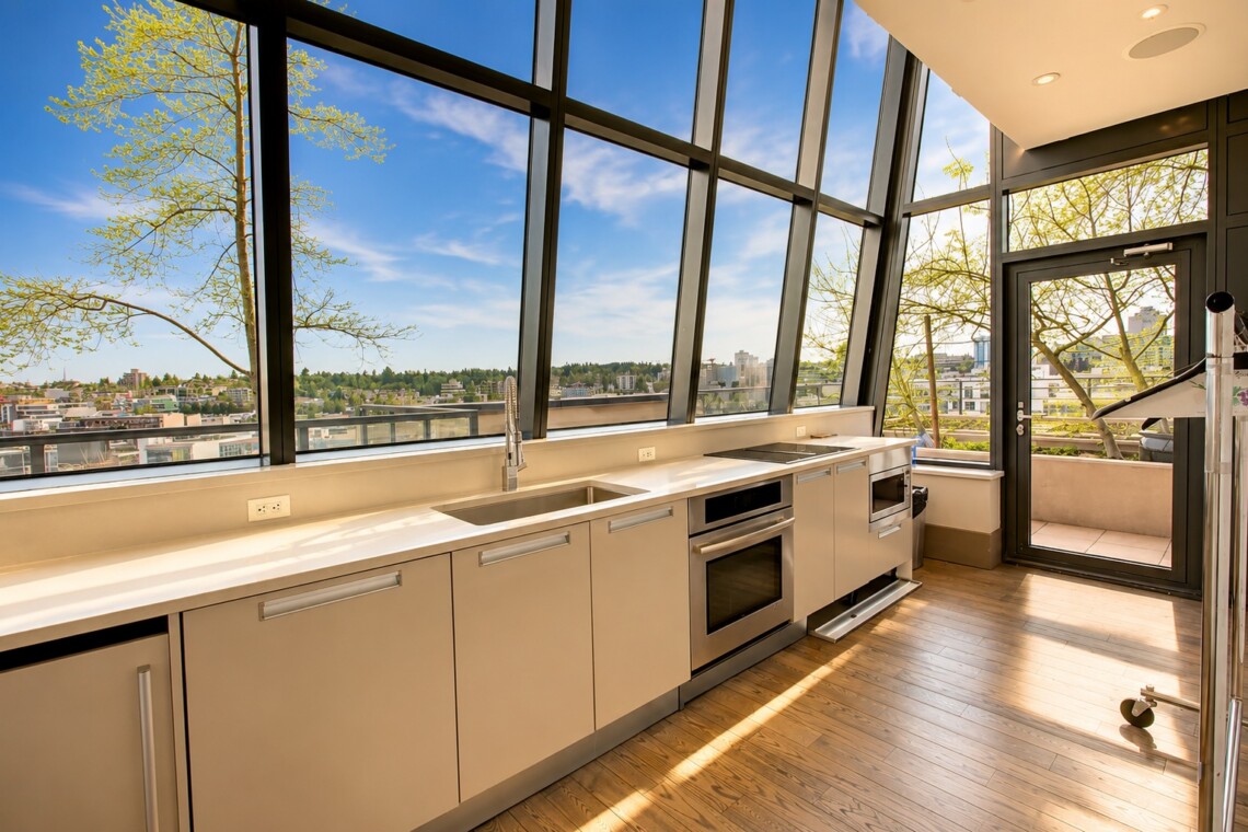 Bright modern kitchen with a long white countertop, built-in oven, sink, and induction hob, set against large angled windows showing a city view and trees outside.