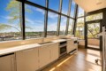 Bright modern kitchen with a long white countertop, built-in oven, sink, and induction hob, set against large angled windows showing a city view and trees outside.