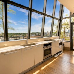 Bright modern kitchen with a long white countertop, built-in oven, sink, and induction hob, set against large angled windows showing a city view and trees outside.