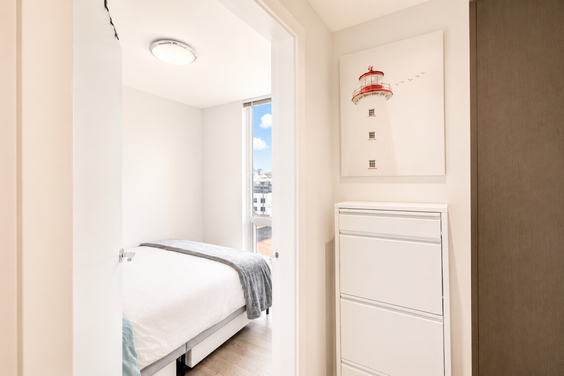 Bright minimalist bedroom with a neatly made white bed, gray throw, and a window showing a city skyline; white dresser on the right and a lighthouse wall art above it.