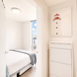Bright minimalist bedroom with a neatly made white bed, gray throw, and a window showing a city skyline; white dresser on the right and a lighthouse wall art above it.
