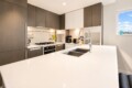 Modern L-shaped kitchen with white quartz island, double stainless sink, and high-arc faucet; dark wood cabinets and a window view to the right.