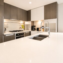 Modern L-shaped kitchen with white quartz island, double stainless sink, and high-arc faucet; dark wood cabinets and a window view to the right.