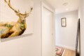 White hallway with a large colorful deer painting on the left wall, a white door in the center, and a small framed print on the far wall; a leaning mirror to the right and a patterned rug on the wooden floor.