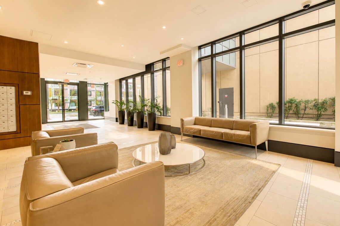 Modern hotel lobby with beige leather sofas, a round glass coffee table, and large floor-to-ceiling windows with potted plants along the wall.