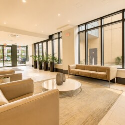 Modern hotel lobby with beige leather sofas, a round glass coffee table, and large floor-to-ceiling windows with potted plants along the wall.