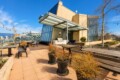 Rooftop patio with wooden tables and benches, string lights, and a glass-walled building against a blue sky, overlooking the city.