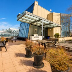 Rooftop patio with wooden tables and benches, string lights, and a glass-walled building against a blue sky, overlooking the city.