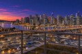 Dusk city skyline over a marina with illuminated high-rise buildings and reflections on the water, seen from a balcony railing in the foreground.
