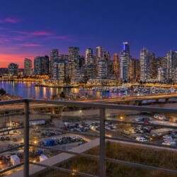 Dusk city skyline over a marina with illuminated high-rise buildings and reflections on the water, seen from a balcony railing in the foreground.