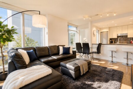 Open-plan living area with a black leather sectional, white fur throw, blue pillows, and a glass dining table near a modern kitchen.