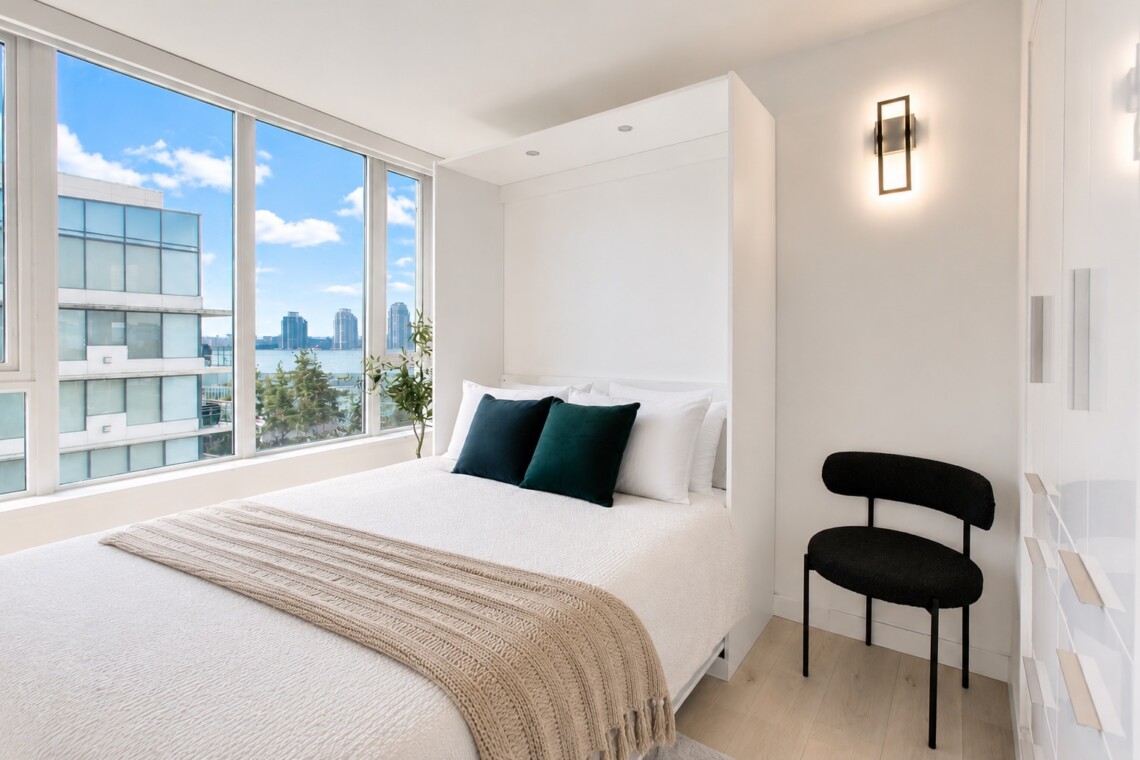 Bright minimalist bedroom with a white bed, beige throw, and two dark green pillows under large windows showing a water and city view.