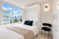 Bright minimalist bedroom with a white bed, beige throw, and two dark green pillows under large windows showing a water and city view.