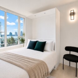 Bright minimalist bedroom with a white bed, beige throw, and two dark green pillows under large windows showing a water and city view.