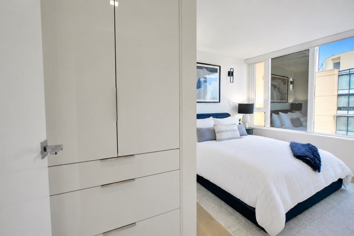 Modern white bedroom with a built-in wardrobe on the left, a bed with white linens and blue pillows, and large windows.