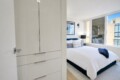 Modern white bedroom with a built-in wardrobe on the left, a bed with white linens and blue pillows, and large windows.