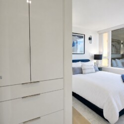 Modern white bedroom with a built-in wardrobe on the left, a bed with white linens and blue pillows, and large windows.