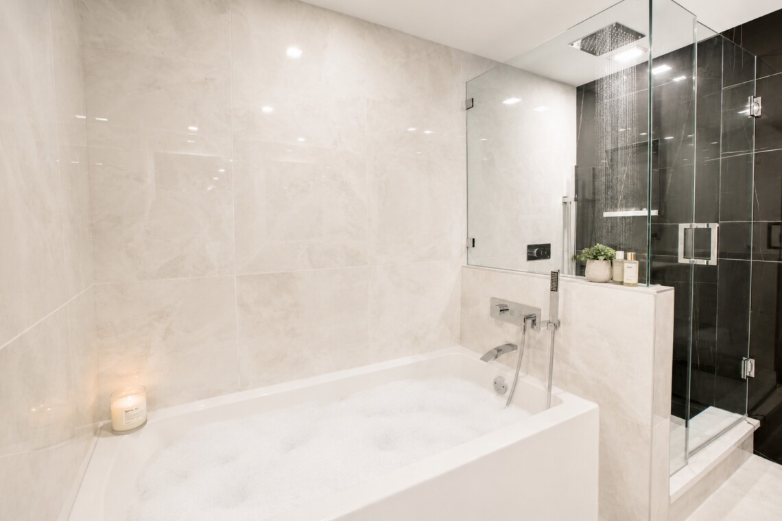 Modern spa-style bathroom with a white built-in tub, beige marble walls, and a glass-enclosed rainfall shower.