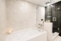 Modern spa-style bathroom with a white built-in tub, beige marble walls, and a glass-enclosed rainfall shower.