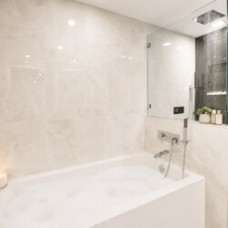 Modern spa-style bathroom with a white built-in tub, beige marble walls, and a glass-enclosed rainfall shower.