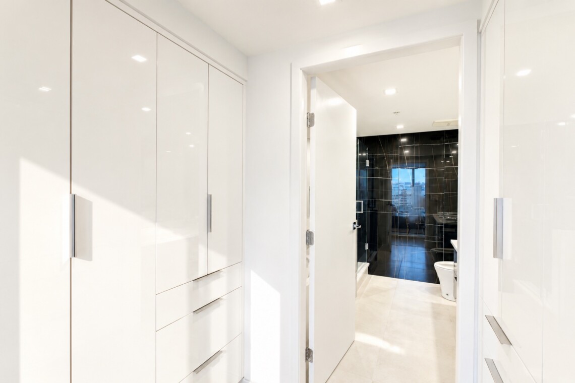 Bright white hallway with built-in wardrobes on the left, leading to a black-tiled bathroom with a glass shower and a city view window in the distance.