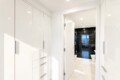 Bright white hallway with built-in wardrobes on the left, leading to a black-tiled bathroom with a glass shower and a city view window in the distance.