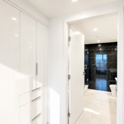 Bright white hallway with built-in wardrobes on the left, leading to a black-tiled bathroom with a glass shower and a city view window in the distance.