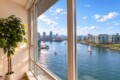 Sunlit room with a large potted plant beside tall windows overlooking a harbor and city skyline at the riverfront.