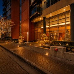Night scene of a modern building entrance adorned with warm Christmas lights on trees and shrubs, seen through large glass doors and windows.