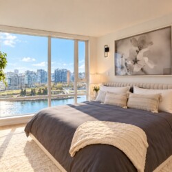 Bright, modern bedroom with a large window overlooking river and city skyline; king bed in dark gray with white and beige pillows and a cream throw.