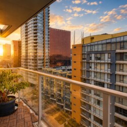 Sunlit cityscape from a balcony, with a potted plant and glass railing at sunset on a high-rise backdrop.