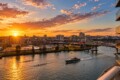 Sunset over a city skyline along a winding river with boats and a nearby bridge.