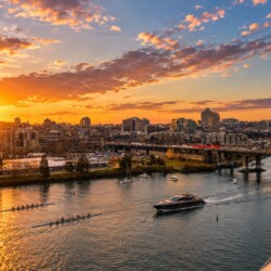 Sunset over a city skyline along a winding river with boats and a nearby bridge.
