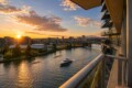 Sunset over a river cityscape viewed from a high balcony, with boats on the water and glass-fronted buildings on the right.