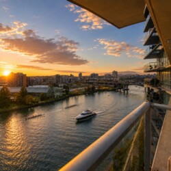 Sunset over a river cityscape viewed from a high balcony, with boats on the water and glass-fronted buildings on the right.