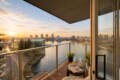 Sunset view from a balcony over a river and city skyline, with a small table set with two coffee cups, a newspaper, and potted plants.