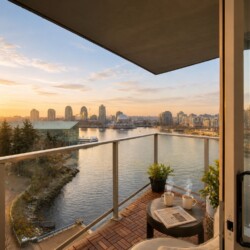 Sunset view from a balcony over a river and city skyline, with a small table set with two coffee cups, a newspaper, and potted plants.