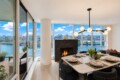 Dining room with a marble table set for six, large windows overlooking a river and cityscape, and a modern chandelier above a lit fireplace.